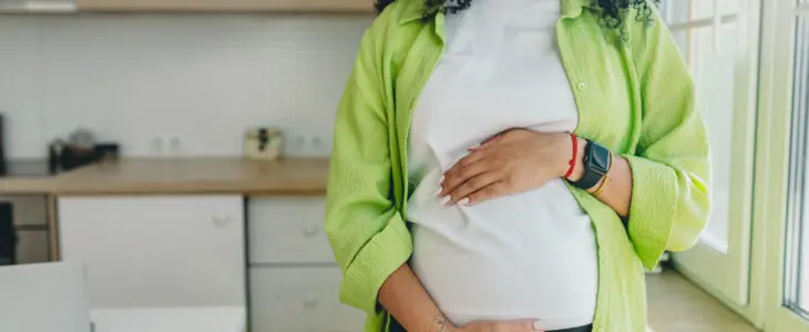 No face picture of pregnant african American woman with long curly hair looking through window standing in kitchen, rubbing her big cute belly, expecting baby boy to be born in few month